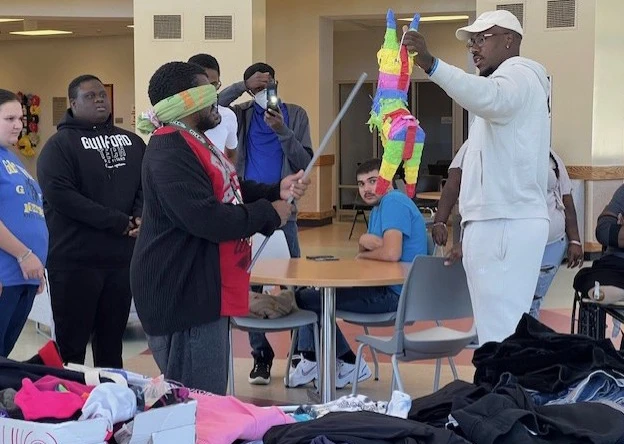 Several male and female members of POWER Pathways watch as a blindfolded male student tries to hit a pinata being held by a male member of GTCC's staff.
