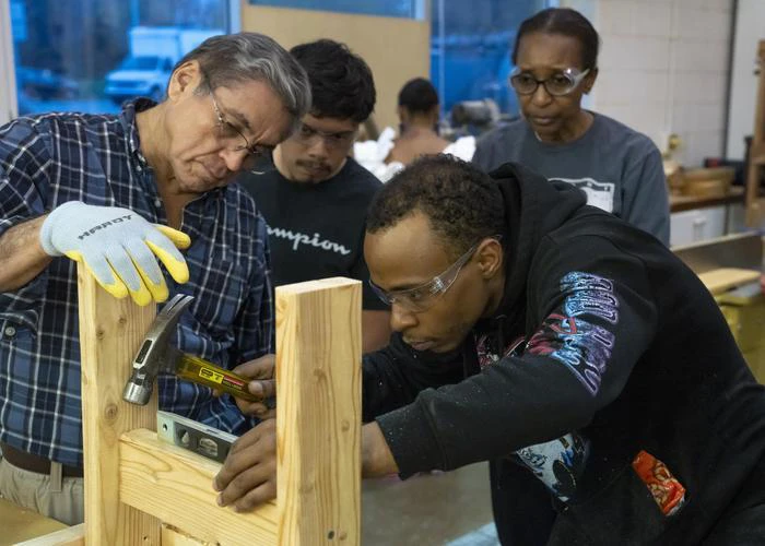 A male instructor works with to male students and a female as they work to level pieces of wood.