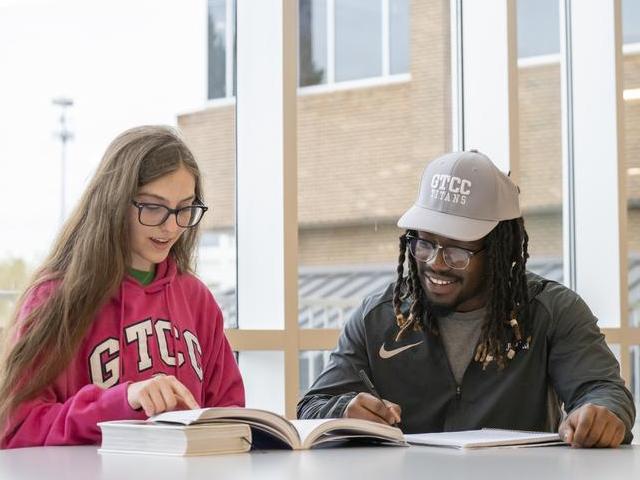 A female and male student sit at a table looking at a book.
