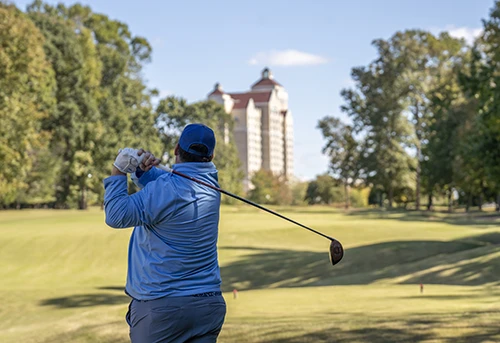 A golf, back to camera, swings with Grandover building in the background.