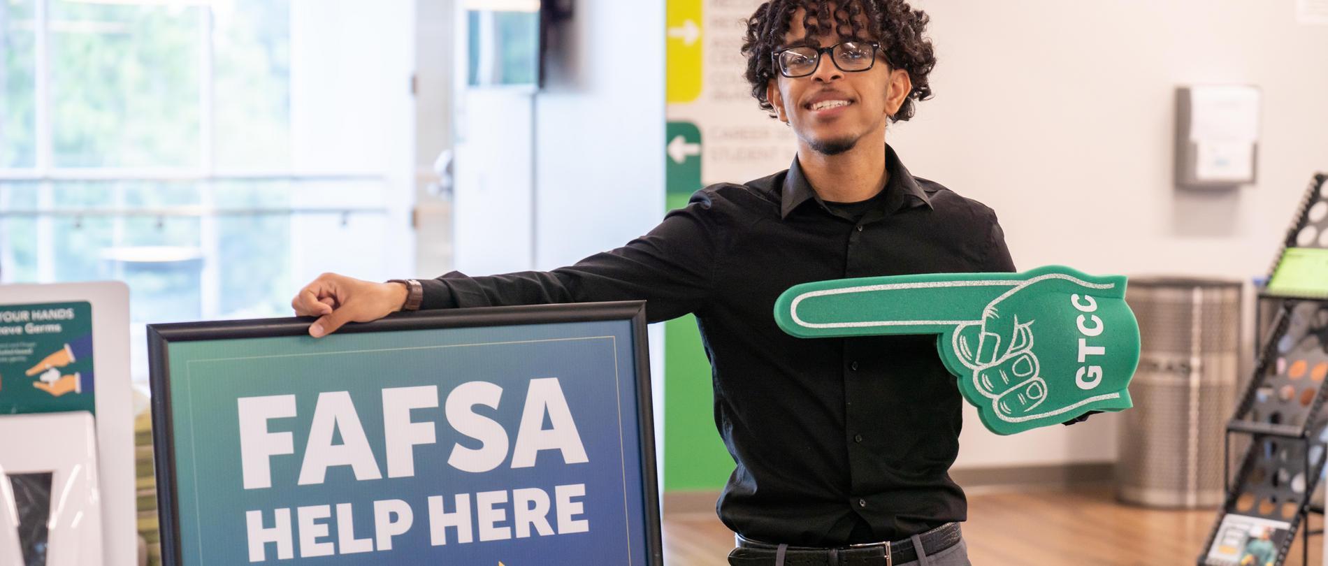 Student smiling while holding a foam finger with “GTCC” on it and a sign that reads “FAFSA Help Here” inside a campus building.