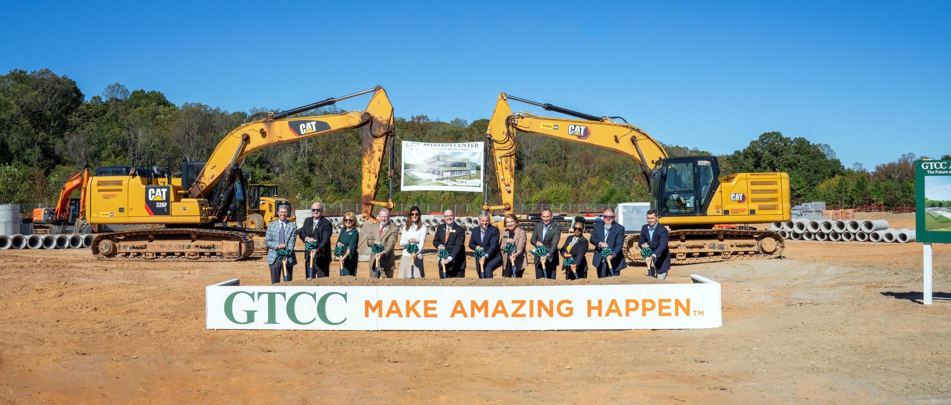 GTCC leaders and partners participate in the Aviation Center groundbreaking ceremony, standing with shovels in front of construction equipment and a banner displaying the future facility rendering.