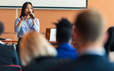 Woman delivers a presentation in front of a large room of people.