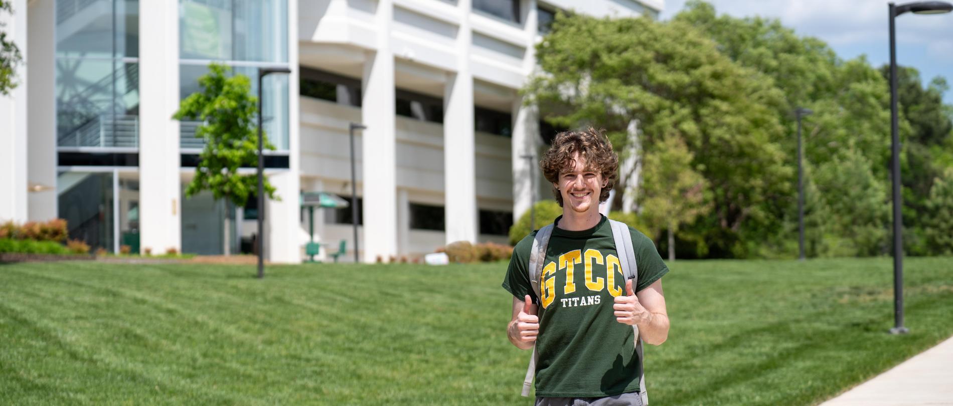 A male student wearing a GTCC T-shirt gives two thumbs up walking on campus in front of a white building.