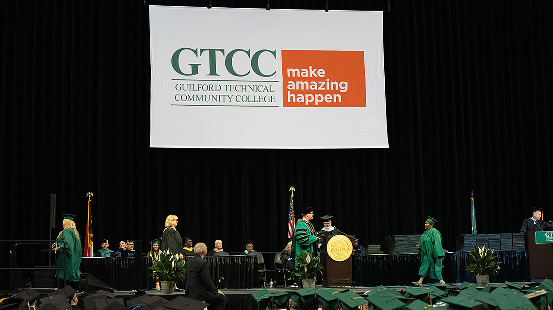 A graduate walks right to left across the stage to receive her diploma.