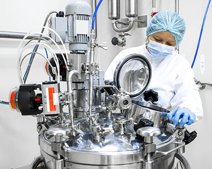 Woman wearing proper protective equipment seen in pharmaceutical manufacturing looking inside the machine that is the part of the drugs production in a pharmaceutical factory.