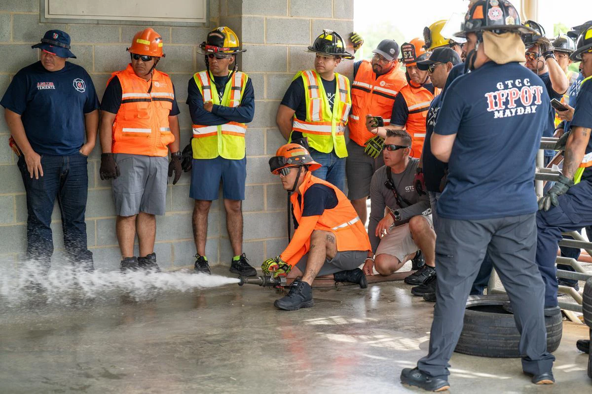 A large group of firefighters watch as one man shoots water out of a hose while kneeling.