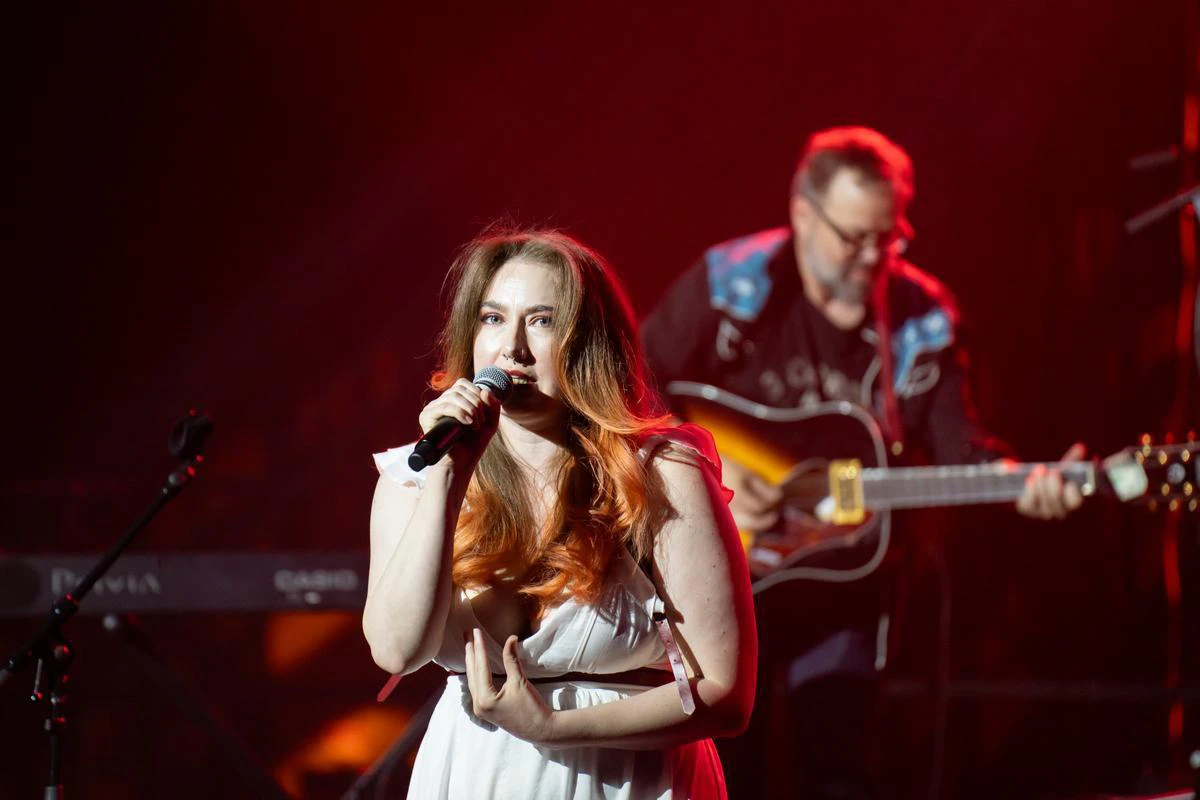 A woman wearing a white dress sings into a microphone with a man wearing a dark jacket playing a guitar in the background.