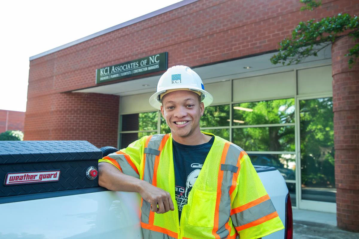 Black male Tsion Mangrum wears a hard hat and yellow reflective vest while leaning on the bed of a white pickup with a brick building in the background.