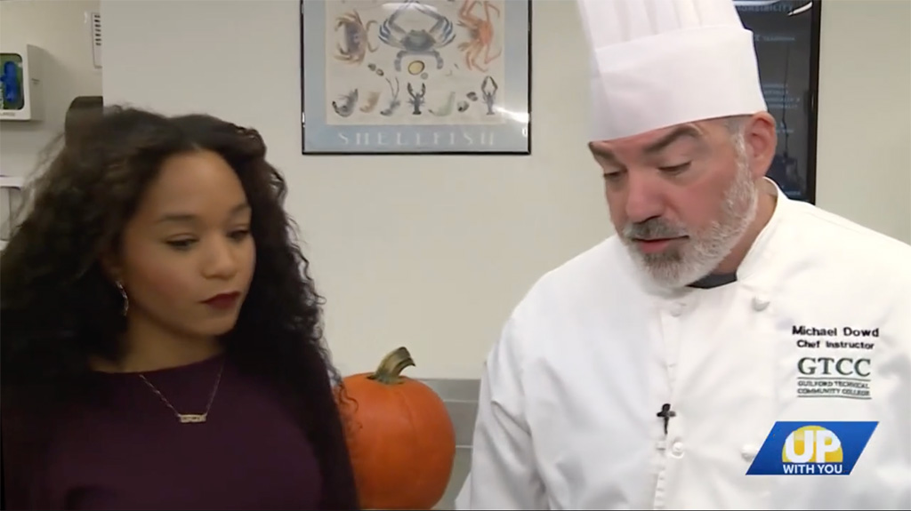 GTCC Chef Instructor Michael Dowd and WXII reporter Erin Burnett stand together in the culinary lab discussing an AI-generated pumpkin cheesecake recipe.