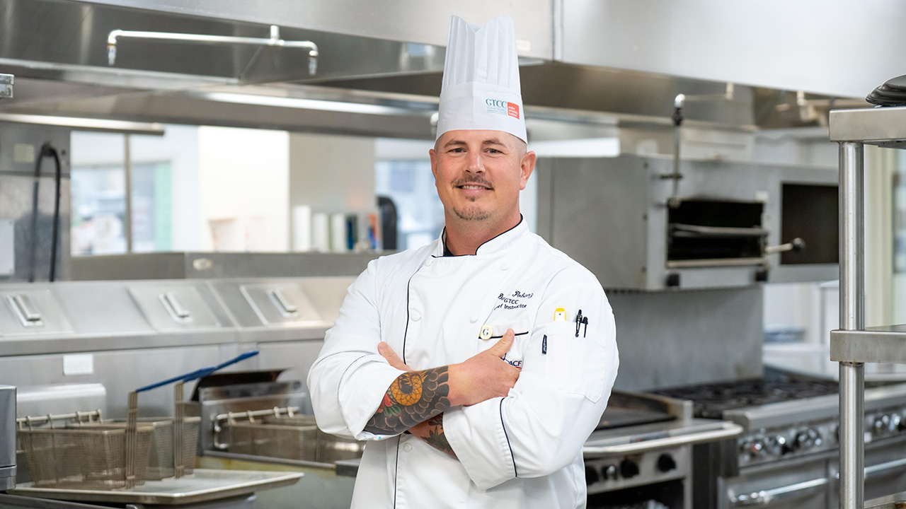 Chef Daryl Pobanz stands confidently in a GTCC culinary lab, wearing his chef’s coat and hat with arms crossed amid professional kitchen equipment.
