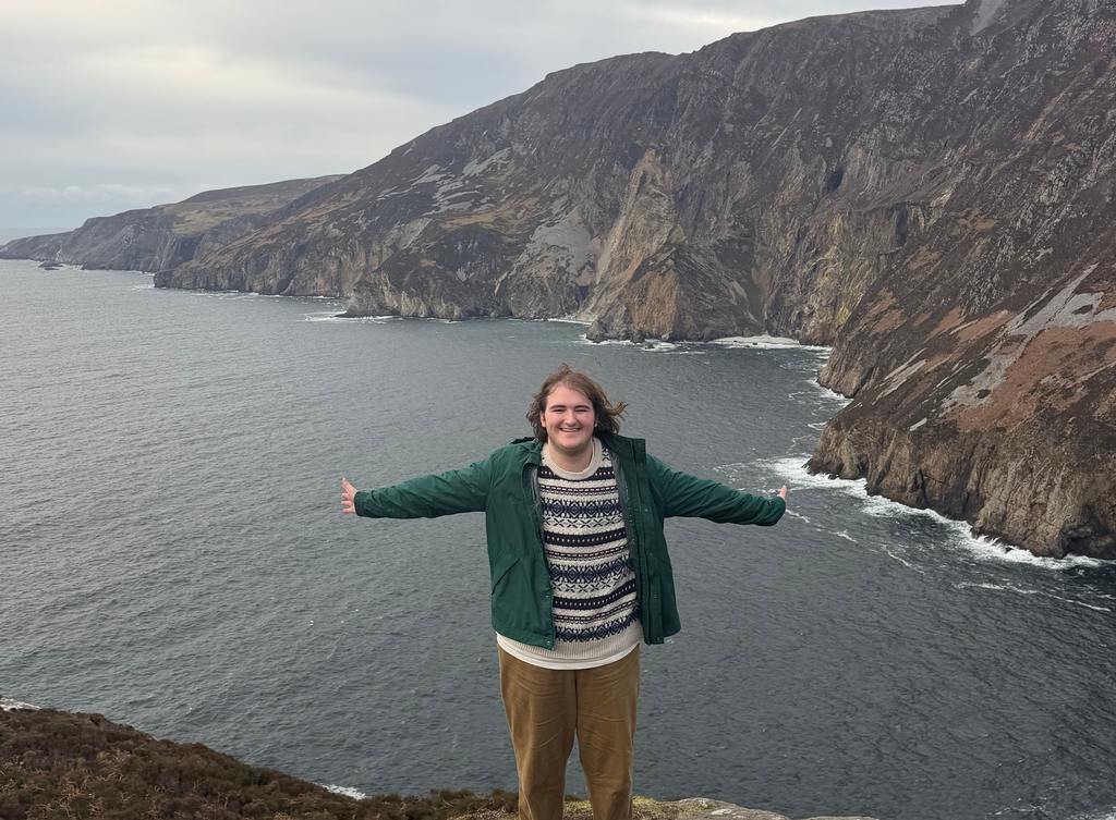 Mason Burghart standing with back to a cliff that overlooks the ocean with a large mountain across the water in the near distance.